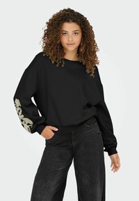 Zwarte oversized sweatshirt met lange mouwen, voorzien van een getextureerd beige bloemenontwerp op de linkerarm. Gecombineerd met losse, donkerdenim jeans.