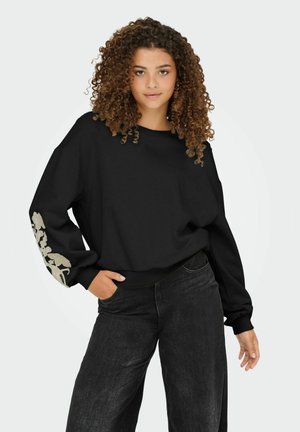 Zwarte oversized sweatshirt met lange mouwen, voorzien van een getextureerd beige bloemenontwerp op de linkerarm. Gecombineerd met losse, donkerdenim jeans.