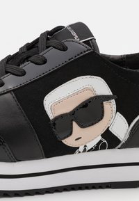 Zapatilla negra de material de ante y cuero, que presenta un personaje de dibujos animados con gafas de sol. Suela gruesa blanca con acentos negros. Cierre de cordones.