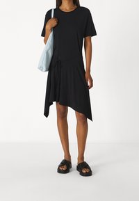 Schwarzes asymmetrisches Kleid mit kurzen Ärmeln, Kordelzug in der Taille, kombiniert mit flachen schwarzen Sandalen und einer hellblauen Tasche. Glatte Stofftextur.