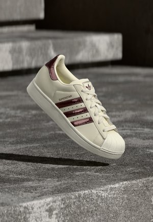 Weißer Adidas Superstar Sneaker mit dunkelroten Streifen und Fersendetail, in der Luft über Betonstufen präsentiert.