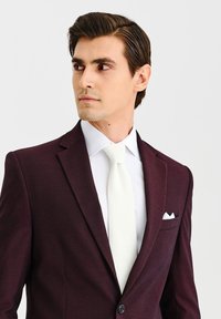 Burgundowa marynarka o slim fit, biała koszula formalna i kołnierzyk w kolorze kości słoniowej. Poszetka w kieszeni piersiowej marynarki.