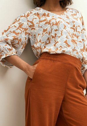 Kvinde iført en løs hvid og rustfarvet mønstret bluse med albuelange ærmer og rustfarvede højttaljede bukser, hånden i lommen, foran en lys væg.