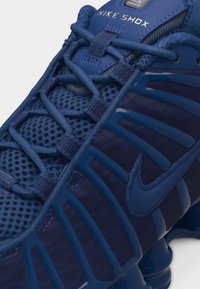 Artimas mėlyno Nike Shox sportbačio vaizdas, kuris rodo tinklinį viršutinį sluoksnį, elastingus raištelius ir būdingą Nike logotipą su amortizuotos pado detalėmis.