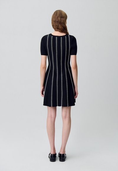 Mujer de pie de espaldas, vistiendo un vestido negro de manga corta con rayas verticales blancas y mocasines negros, sobre un fondo liso.