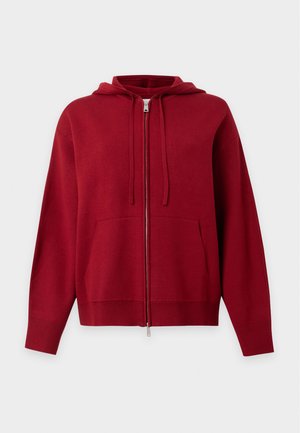Une femme porte un sweat à capuche rouge zippé par-dessus un haut rayé, un legging corsaire noir, une casquette marron et des bottes marron foncé.