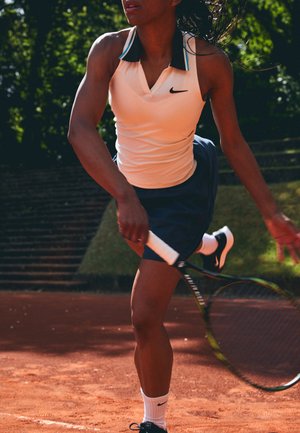 Tennisantrekk som inkluderer en ermeløs hvit topp med svart krage og blå detaljer, kombinert med en marineblå skjørt og svarte joggesko.