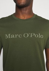 Oliven grøn kortærmet t-shirt lavet af blødt stof med "Marc O'Polo" i lys grå tekst henover brystet. Klassisk crew neck design.