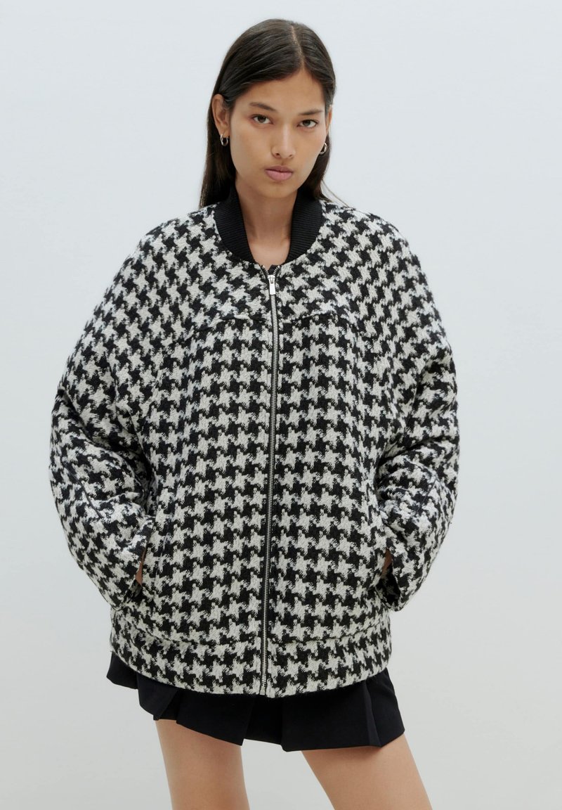 Bomberjack met een houndstooth-patroon in zwart en wit, met een ritssluiting en geribde kraag. Textuurstof met zijzakken.