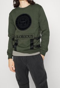 Persoon in een olijfgroene sweatshirt met zwart cirkelvormig embleem en de tekst "Glorious", gecombineerd met donkergrijze cargobroek.
