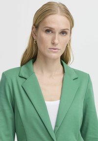 Groene blazer met een gladde textuur, met kraagjes en een getailleerde pasvorm, gecombineerd met een witte top. Bevat subtiele stikseldetails.
