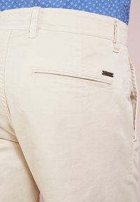 Beiget puuvillahousut, joissa on takatasku piilotetulla vetoketjulla ja pieni metallinen brändäyslevy. Sileä pinta.