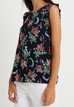 Blouse noire sans manches avec des imprimés floraux et animaliers colorés portée avec un pantalon blanc sur une personne debout.