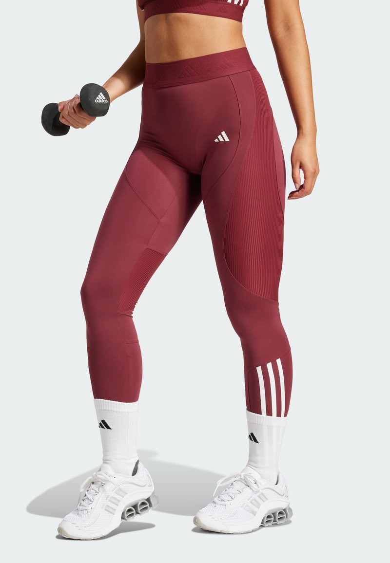 Bordeaux träningsleggings med ribbad textur, med hög midja och accentpaneler. Matchas med vita strumpor och skor, hållande en hantel.