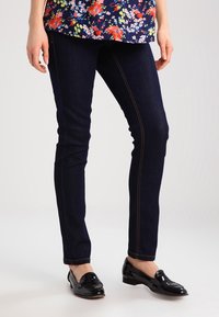 Tmavě modré skinny džíny s kontrastním oranžovým šitím, vyrobené z denimu, v kombinaci s černými lakovanými mokasíny.