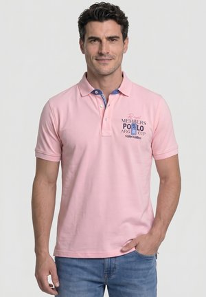 Hombre con camisa polo rosa claro con detalles azules y texto impreso, sonriendo, con una mano en el bolsillo de los vaqueros azules, de pie frente a un fondo liso.