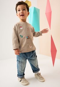 Sweat-shirt beige clair avec broderie colorée sur le thème des fêtes, associé à un jean bleu ample et des baskets crème, posant devant des décors géométriques.