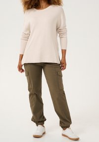 Beiger, gerippter Pullover mit langen Ärmeln, kombiniert mit olivgrünen Cargohosen mit Seitentaschen und elastischen Bündchen, getragen mit weißen Turnschuhen.