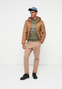 Giubbotto puffer marrone con cappuccio, felpa verde con logo, pantaloni beige e sneakers nere, indossati da una persona in un ambiente neutro.