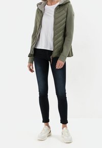 Giacca imbottita verde oliva con cappuccio, chiusura con zip e polsini elasticizzati, abbinata a una maglietta bianca e jeans skinny scuri, completata da sneakers bianche.