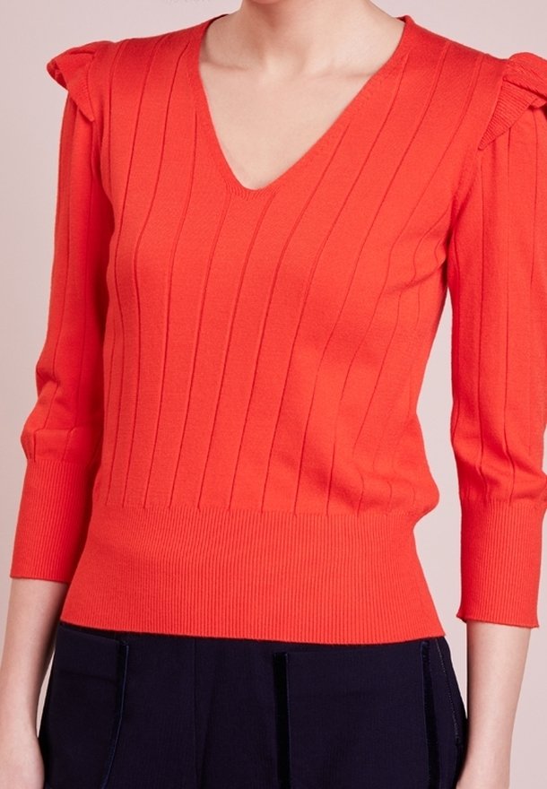 Femme portant un pull orange vif à col en V avec côtes verticales et manches 3/4, associé à un pantalon foncé.
