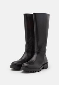 Botas altas de cuero negro con acabado brillante, puntera redonda y suela de goma gruesa con surcos profundos para mayor tracción.