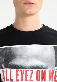 T-shirt en coton noir avec un grand imprimé graphique d'un visage en noir et blanc, accompagné d'un texte rouge indiquant "TOUS LES YEUX SUR MOI" en dessous.