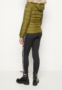 Oliven grøn pufferjakke med pelsforet hætte, kombineret med sorte leggings med en hvid logo-stribe og sorte snørebånds støvler.