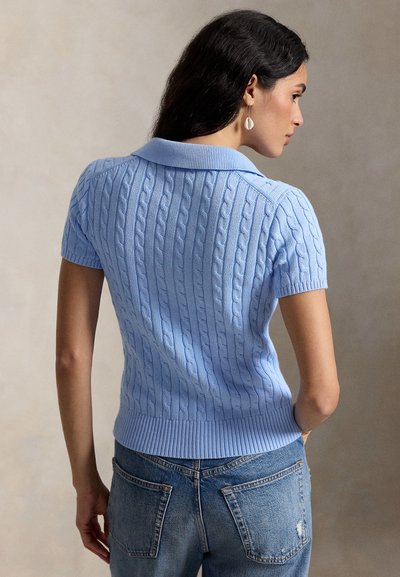 Femeie cu păr închis la culoare, purtând un pulover cu mânecă scurtă, tricotat cu model torsadat, de culoare albastru deschis, și blugi albaștri, văzută din spate și profil lateral.