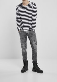 Camicia a maniche lunghe a righe bianche e nere, jeans grigi slim-fit e anfibi neri con lacci e finitura testurizzata. Il modello è posizionato su uno sfondo neutro.