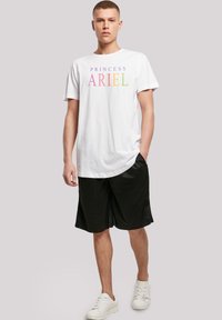 White cotton T-shirt with rainbow text "PRINCESS ARIEL" on the front. Paired with black athletic shorts and white sneakers. Standing posture.