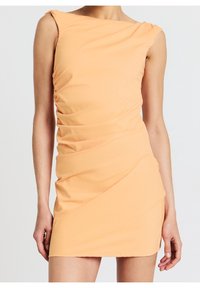 Mini-robe ajustée sans manches couleur pêche-orange avec fronces sur les côtés portée par une personne debout devant un fond uni.