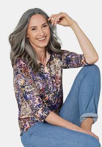 Mehrfarbige Bluse mit floralem und abstraktem Muster, mit Knopfleiste, Dreiviertelärmeln, kombiniert mit hellblauen Jeans.