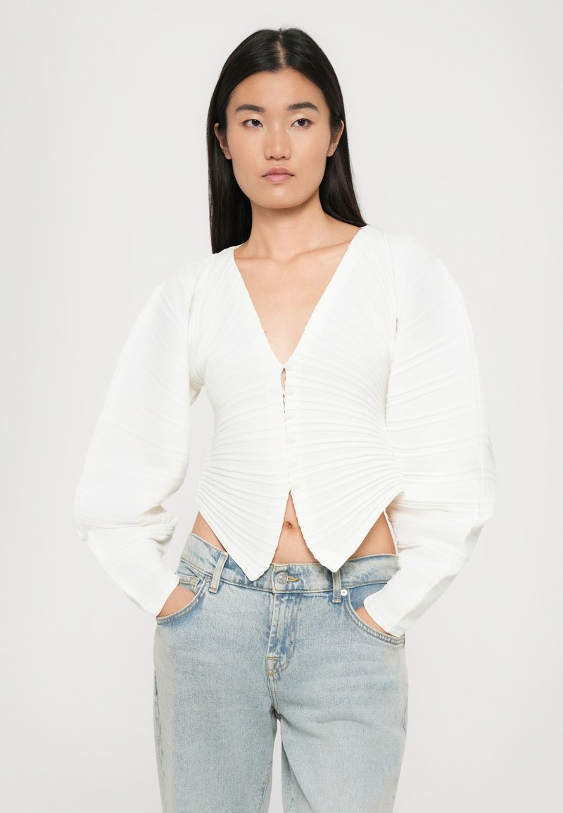 Haut blanc ajusté avec un tissu texturé et plissé, décolleté en V profond et manches longues bouffantes. Doté de boutons sur le devant et d'un design raccourci.
