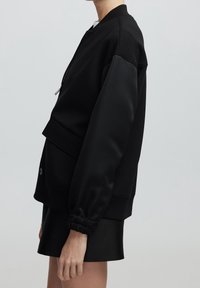 Zwarte bomberjack met een rits aan de voorkant, elastische mouwen en een gelaagd ontwerp. Gemaakt van een gladde stof met een matte afwerking.