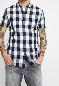 Camicia a maniche corte con bottoni in pattern a quadri blu navy e bianco, dotata di taschino sul petto e dettagli del colletto a contrasto, realizzata in tessuto a trama.