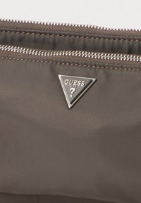 Grå nylonpose med sølvglidelås, med et trekantet metalllogo som leser "GUESS?" nær glidelåsen.