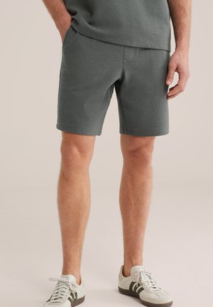 Mees, kes kannab halli tekstuuriga lühike pükse ja valgeid tossusid, seisab ühe käega taskus neutraalse tausta ees.