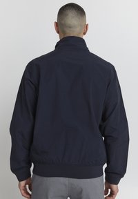Tamno plavi vjetrovka s visokim ovratnikom, elastičnim manžetama i pojasom. Glatka tekstura, minimalistički dizajn, bez vidljivih logotipa ili uzoraka.