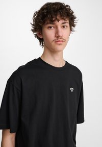 Camiseta de algodón negra con cuello redondo y mangas cortas, que presenta un pequeño logotipo en la parte izquierda del pecho. Textura suave.