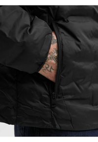 Schwarze Pufferjacke mit einer Reißverschlusstasche, die eine glatte Textur und ein gesteppter Design zeigt. Das Material scheint leicht und wasserabweisend zu sein.