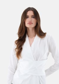 Blusa bianca avvolgente con scollatura a V, maniche lunghe e un elegante dettaglio in vita a strati. Tessuto dalla consistenza liscia su uno sfondo chiaro.