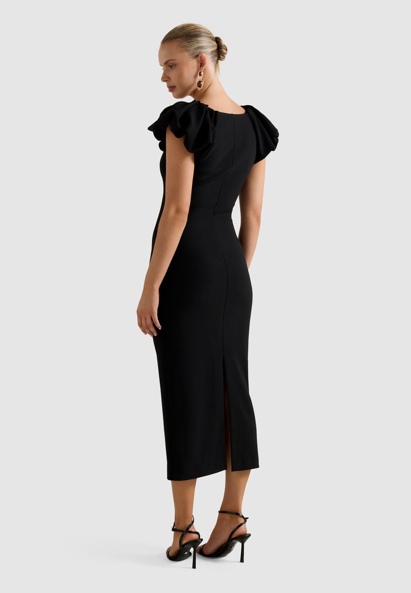 Vrouw in een getailleerde zwarte midi-jurk met ruches op de schouders, een split aan de achterkant, zwarte sandalen met hoge hakken en gouden hoepel oorbellen, staand in de zijaanzicht.