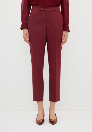 Burgundy tailored trousers with a smooth texture, featuring a button closure, pleated front, and cropped length. Paired with pointed-toe shoes.