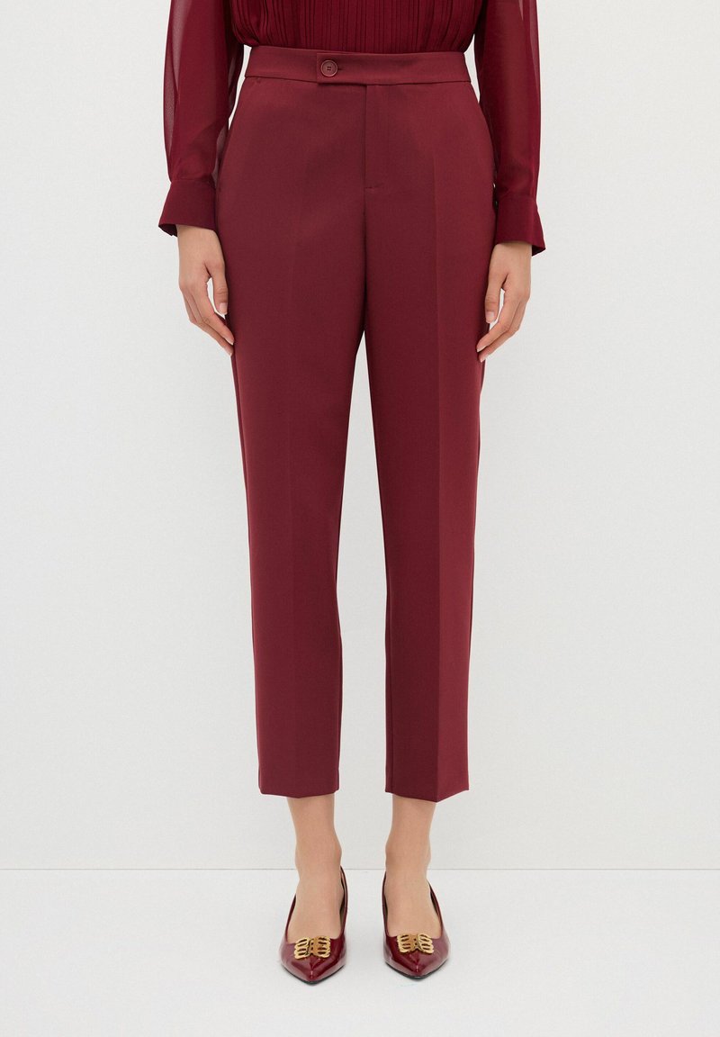 Pantaloni su misura color borgogna con una texture liscia, dotati di chiusura a bottone, frontale plissè e lunghezza croppata. Abbinati a scarpe con punta affilata.