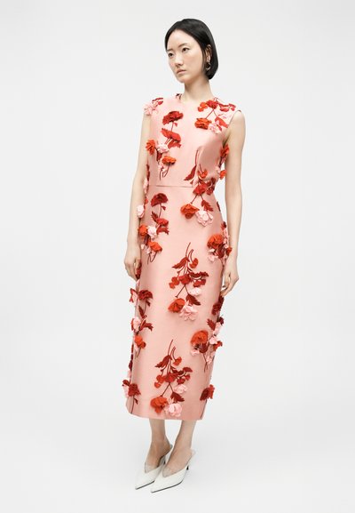 Naine, kes kannab varrukateta roosat midi-kleiti, millel on tekstureeritud punaseid ja roosasid lillelisandusi, koos valgete teravate ninaga kontsakingadega.