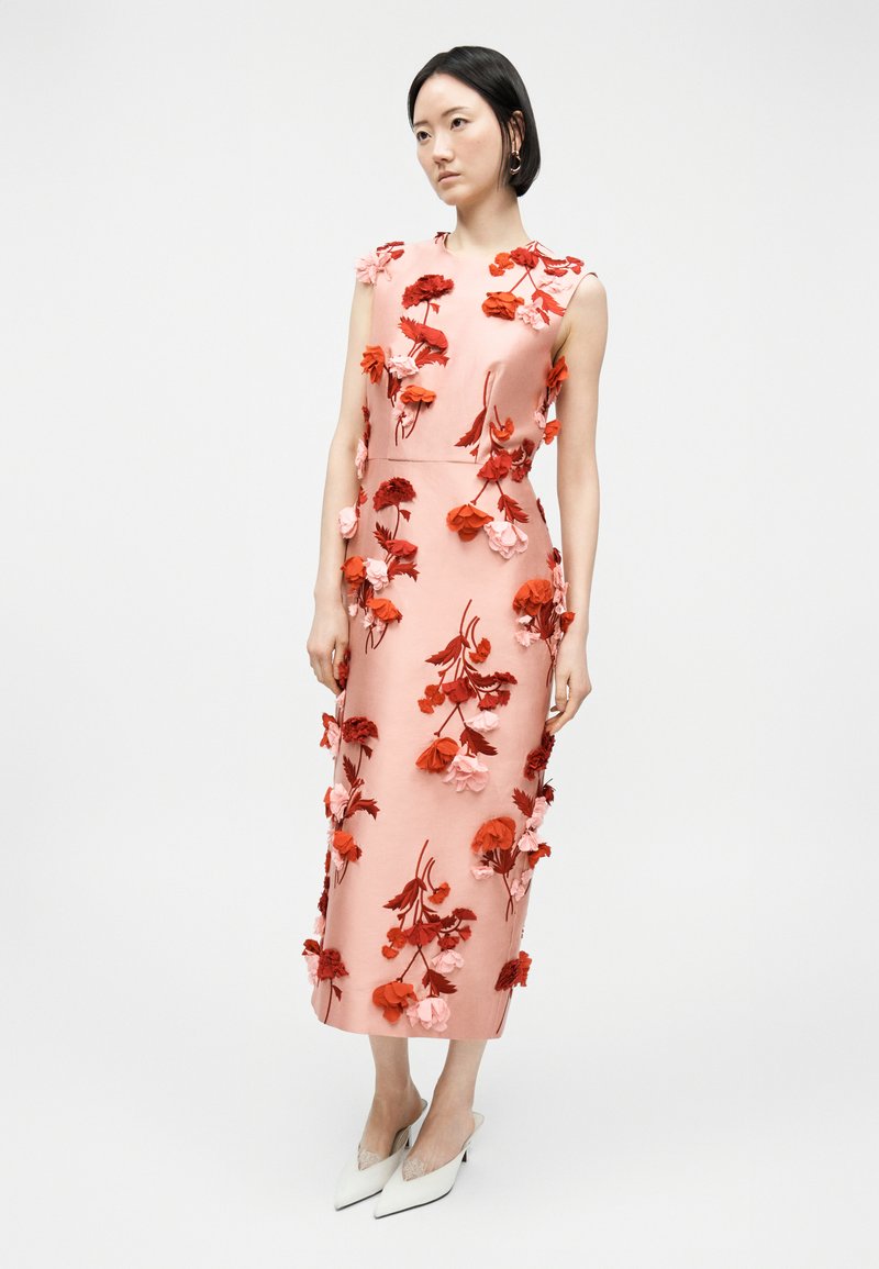 Donna che indossa un abito midi rosa senza maniche con applicazioni floreali rosse e rosa testurizzate, abbinato a décolleté bianchi con punta a punta.
