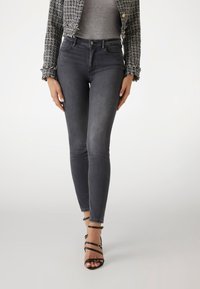 Slanke grijze jeans met een hoge taille, voorzien van een zachte textuur en subtiele vervaging. Draagt met zwarte sandalen met banden en een gestructureerde zwarte jas.