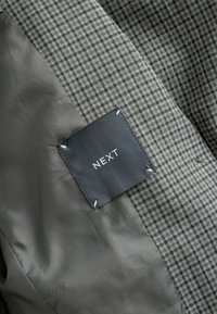 Hall ja musta huntstooth mustriga kangas, sileda halli voodriga; sisaldab musta silti, kus on valge tekstiga "NEXT".