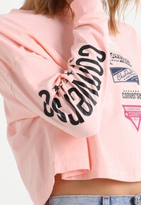 Ljusrosa kortärmad topp med långa ärmar med svart tryckt text och grafik på framsidan och ärmen. Tillverkad av mjukt bomullsmaterial.